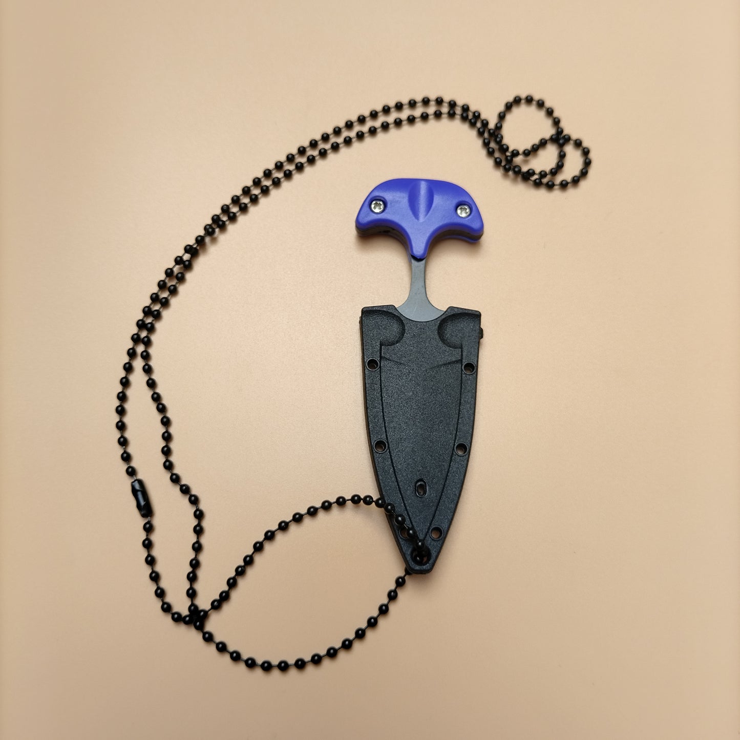 A black neck knife with a sheath and neck chain, displayed on a wooden surface with pine cones and a leaf in the background.