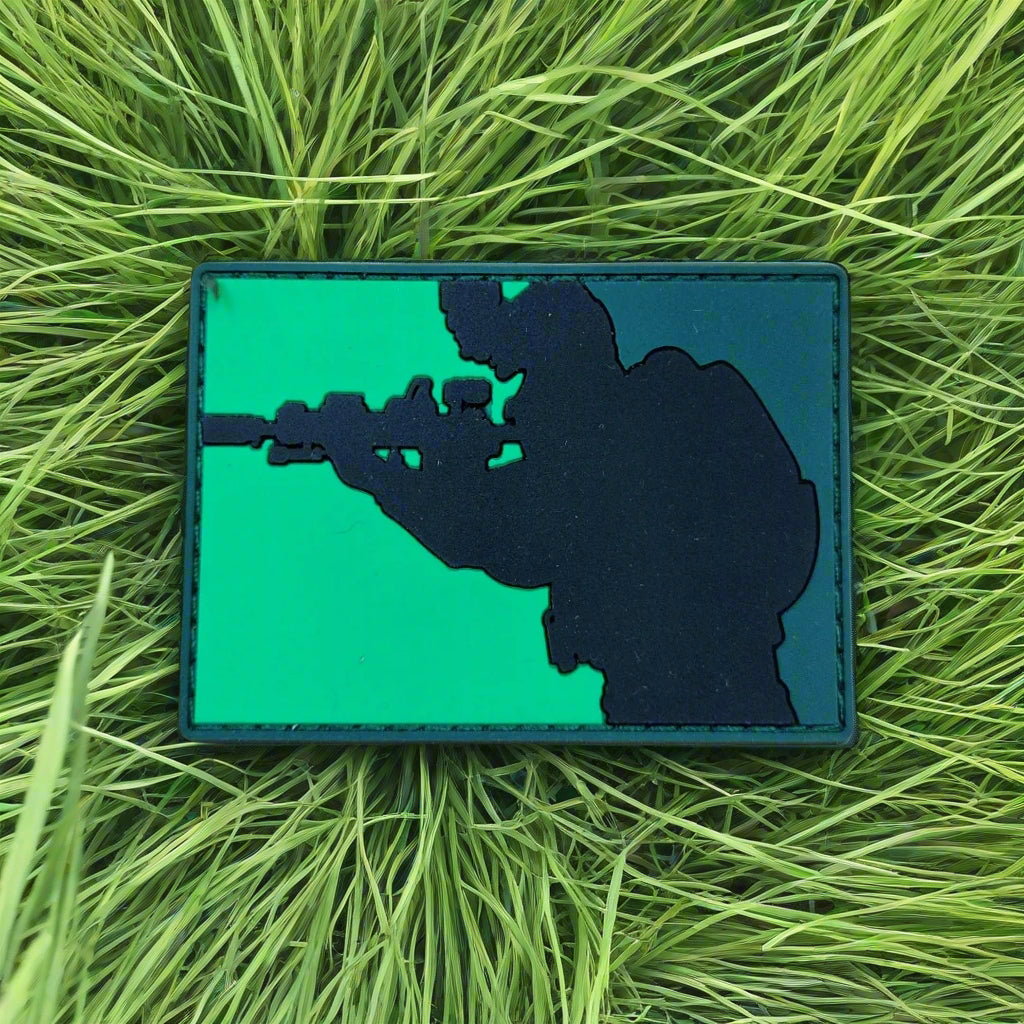 Silhouette patch of a person with a gun on a green background, lying on grass.