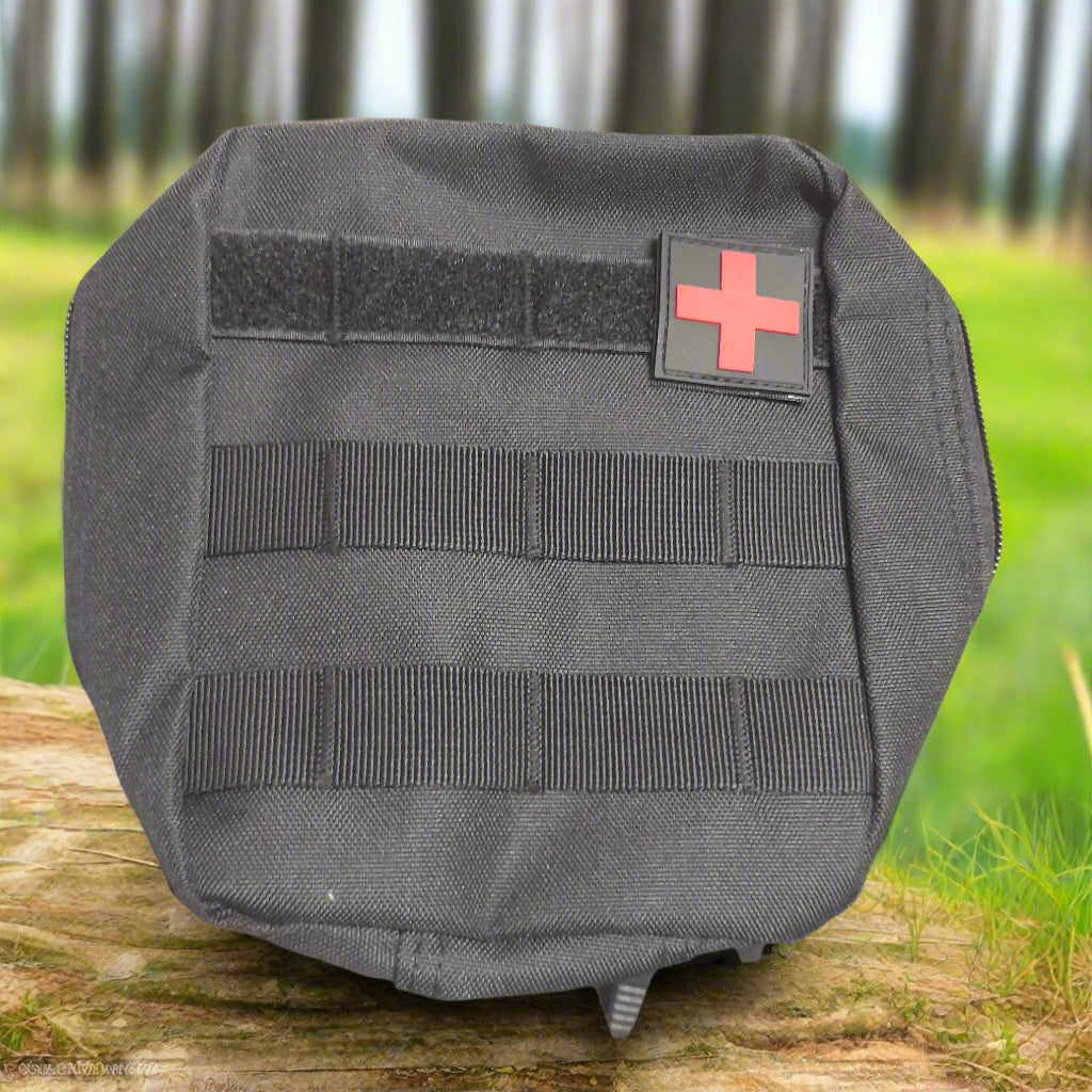 Black tactical pouch with a red cross patch on a wooden surface with grass and trees in the background