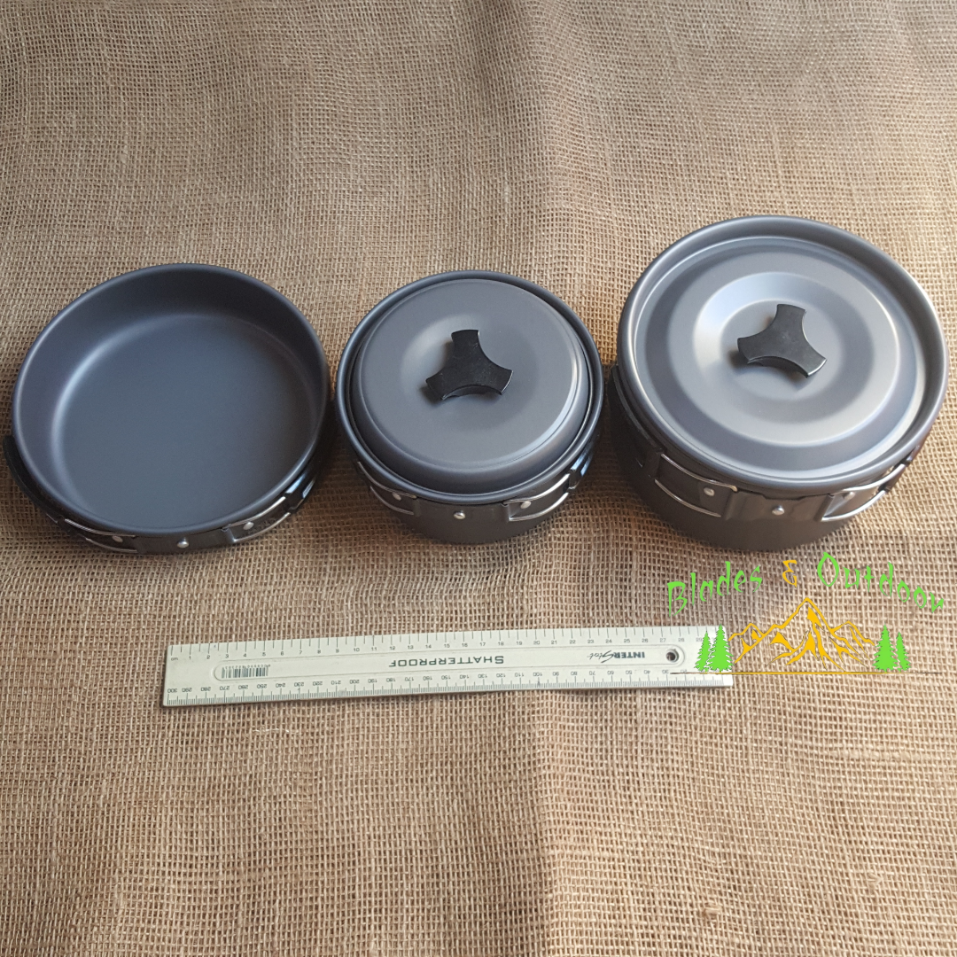 Set of camping cookware including pots, pans, and utensils on a wooden board with a natural outdoor background.