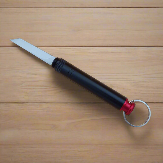 A black keyring whistle knife with a hidden blade, placed next to its neat plastic box on a beige background.