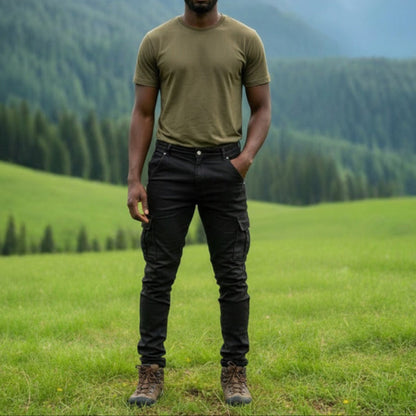 Man wearing black 6 pocket jeans, standing in a grassy field with mountains in the background. 
