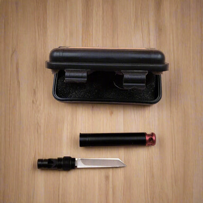 A black keyring whistle knife with a hidden blade, placed next to its neat plastic box on a beige background.