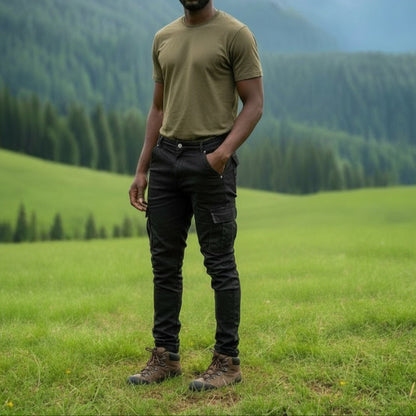 Man wearing black 6 pocket jeans, standing in a grassy field with mountains in the background. 