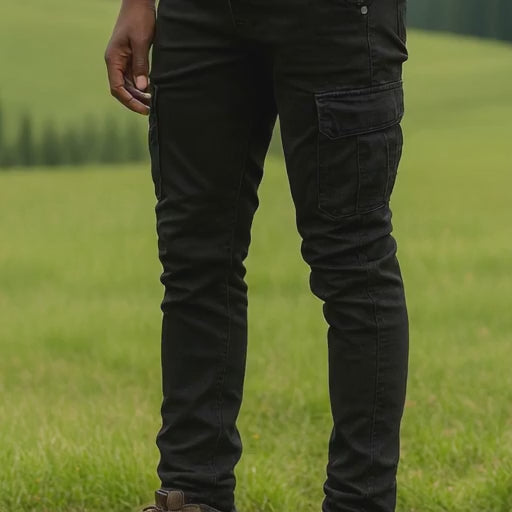 Man wearing black 6 pocket jeans, standing in a grassy field with mountains in the background. 