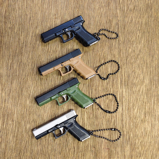 Four miniature handgun-shaped keychains with black ball chains, displayed on a light brown wood grain surface. The keychains are shown in four color options: matte black, tan, olive green, and silver and black.
