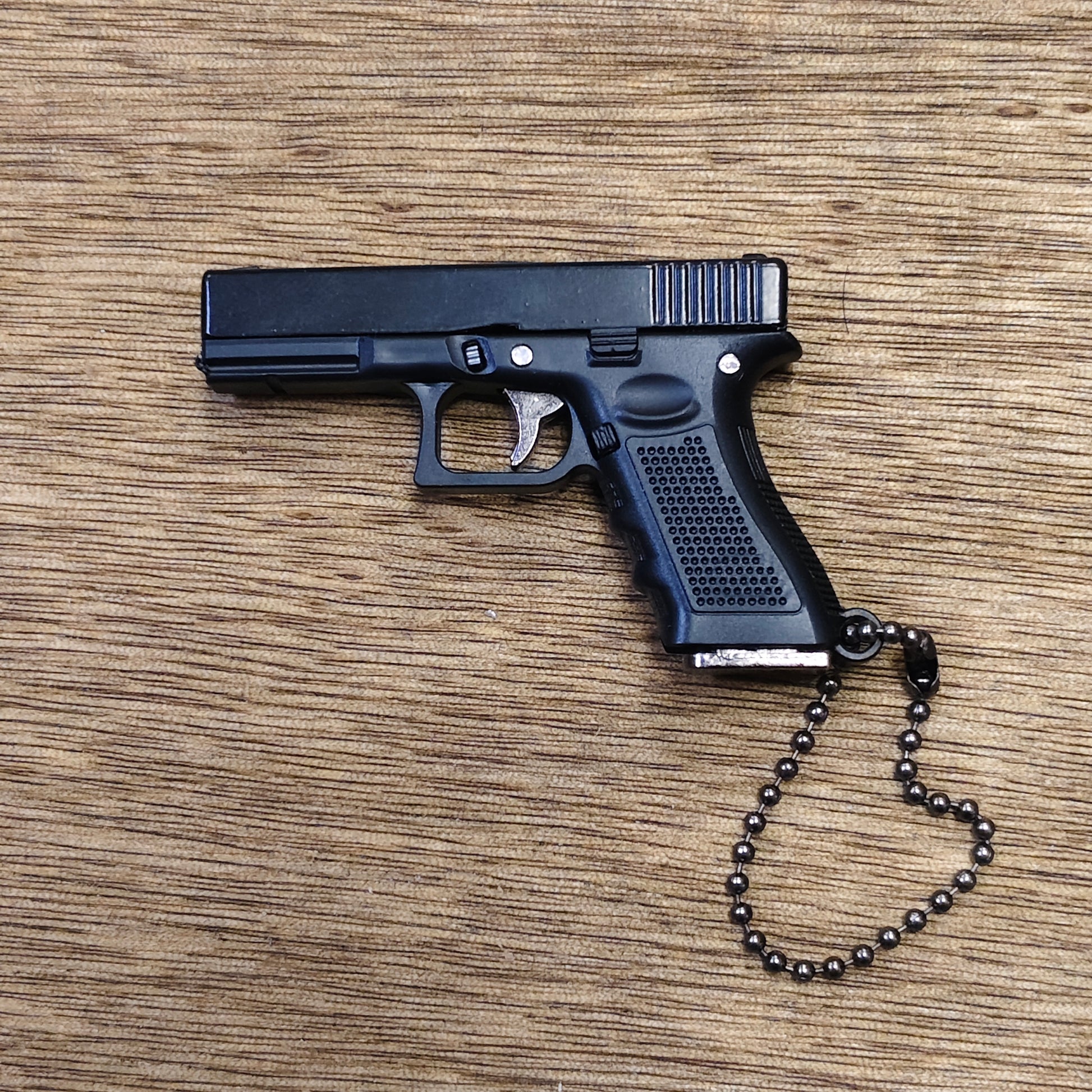 A black replica semi-automatic keyring pistol, styled like a Glock model, resting on a brown wood-grain surface. It features a black ball chain attached to the grip.