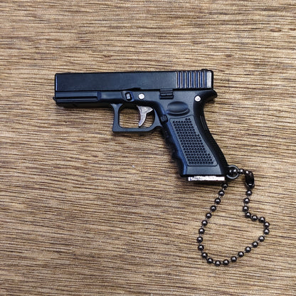 A black replica semi-automatic keyring pistol, styled like a Glock model, resting on a brown wood-grain surface. It features a black ball chain attached to the grip.