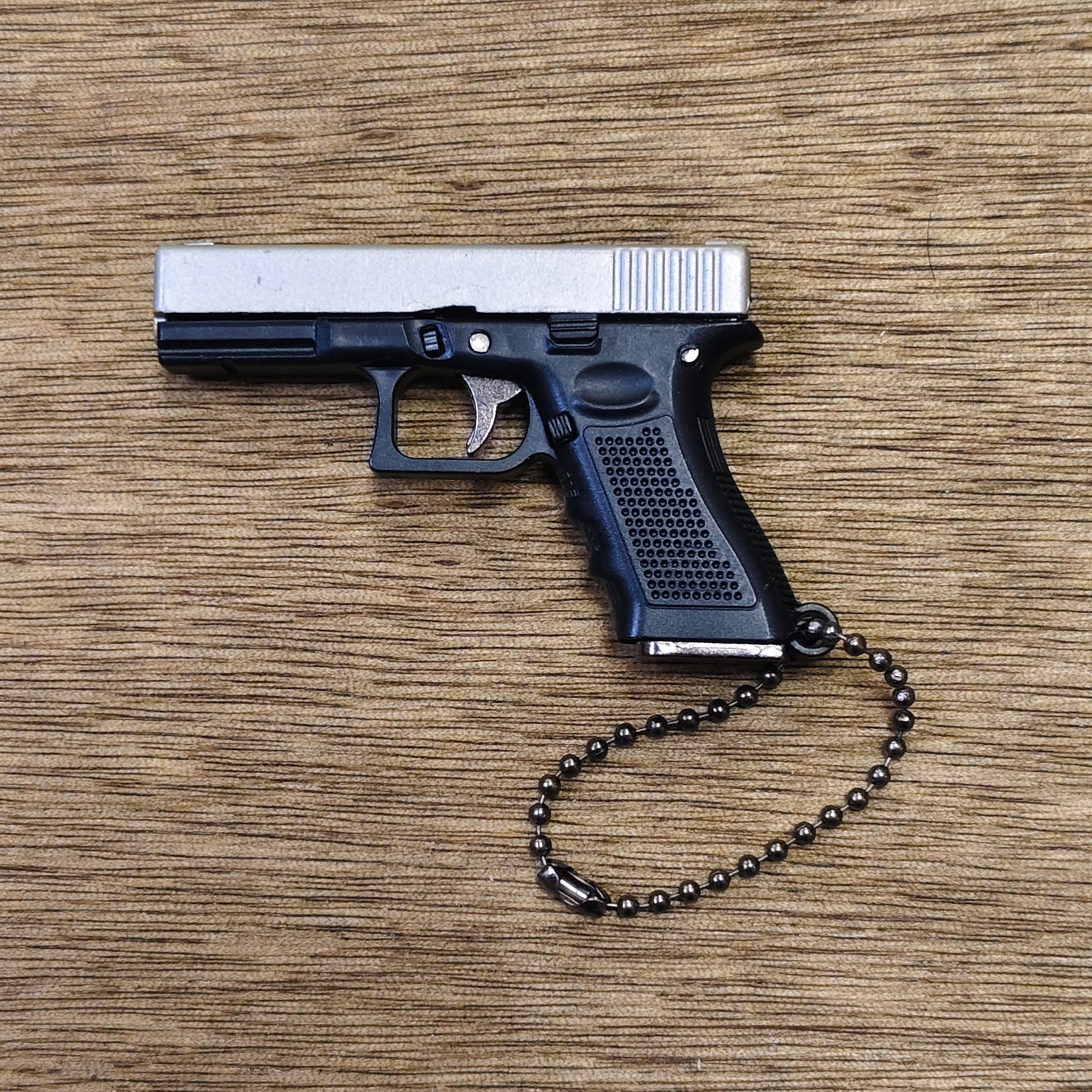 A miniature replica pistol keyring, styled like a semi-automatic handgun, with a silver slide and black grip, attached to a black beaded keychain, displayed on a wooden surface.