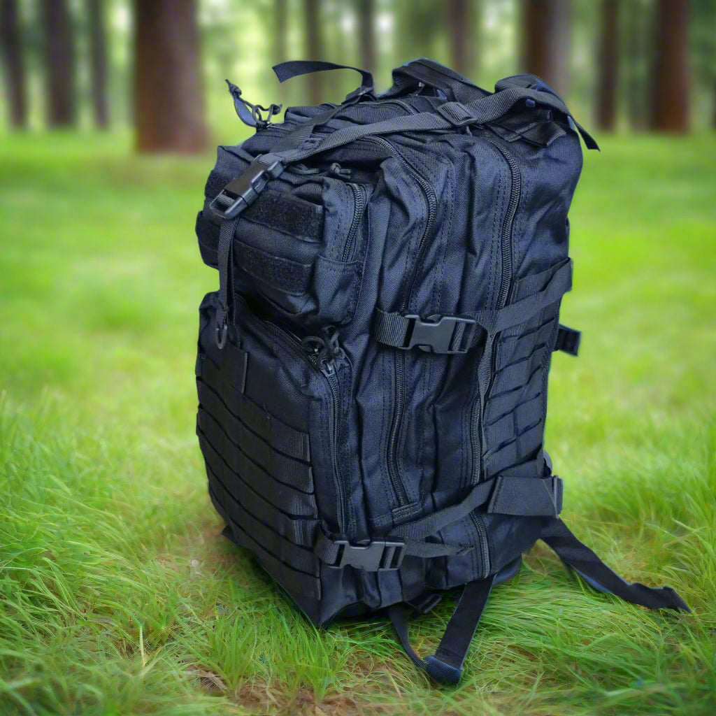 Black backpack surrounded by green grass, in a forest, mountains, and trees in the background