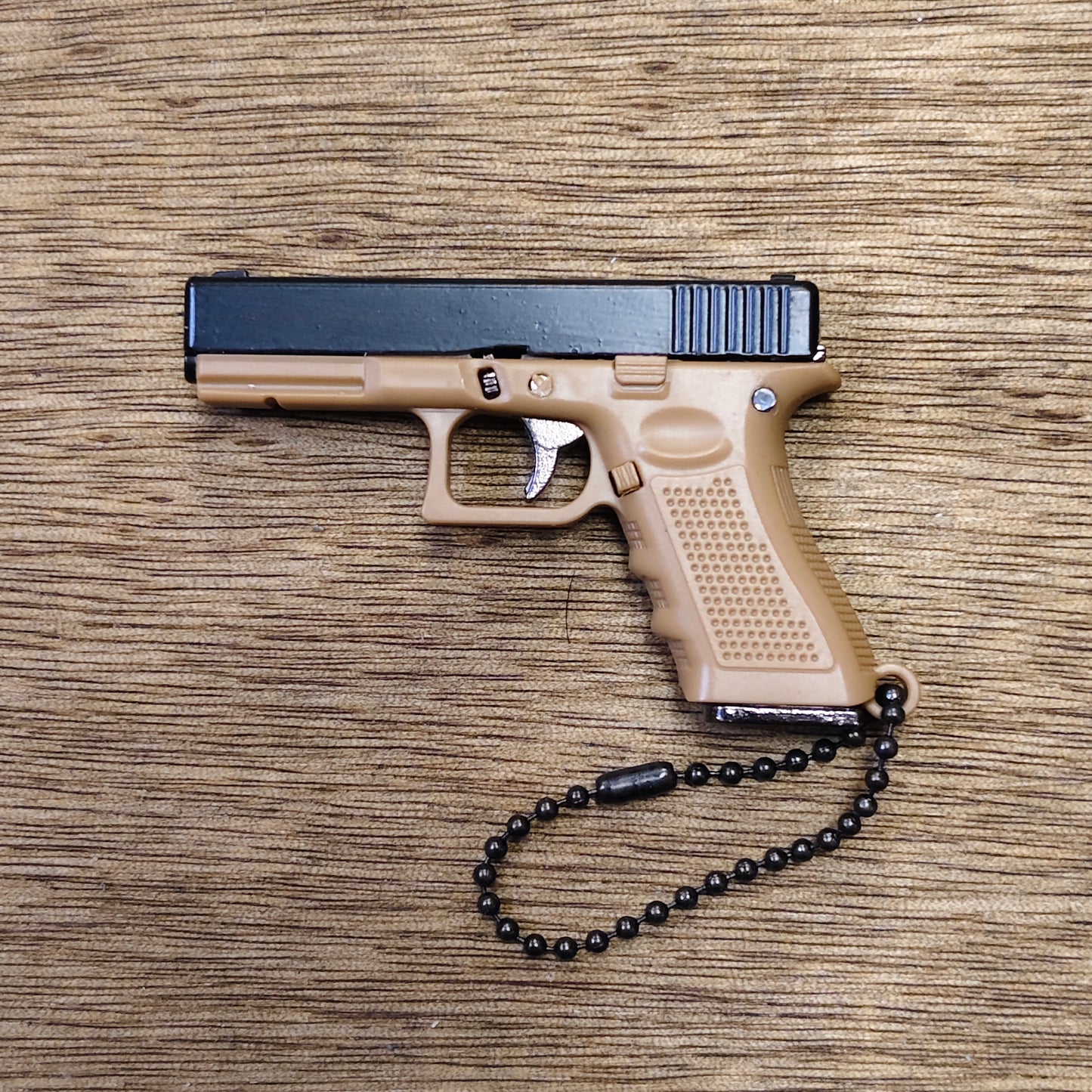 A compact semi-automatic keyring pistol in tan and black, photographed from above on a wood-grain surface, with a black beaded wrist chain attached to the grip.