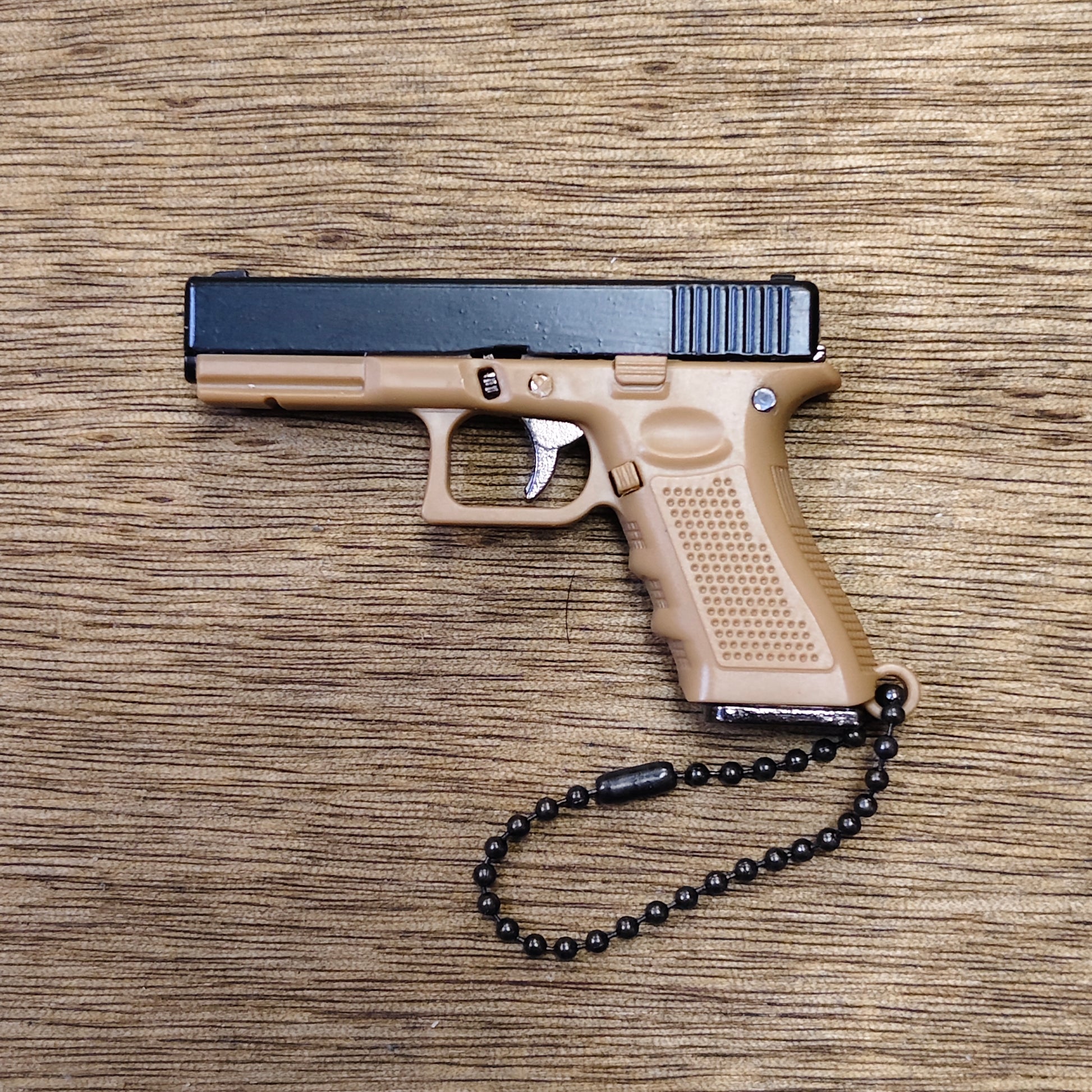 A compact semi-automatic keyring pistol in tan and black, photographed from above on a wood-grain surface, with a black beaded wrist chain attached to the grip.