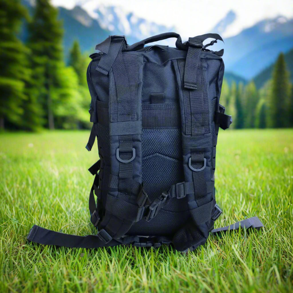 Black backpack surrounded by green grass, in a forest, mountains, and trees in the background