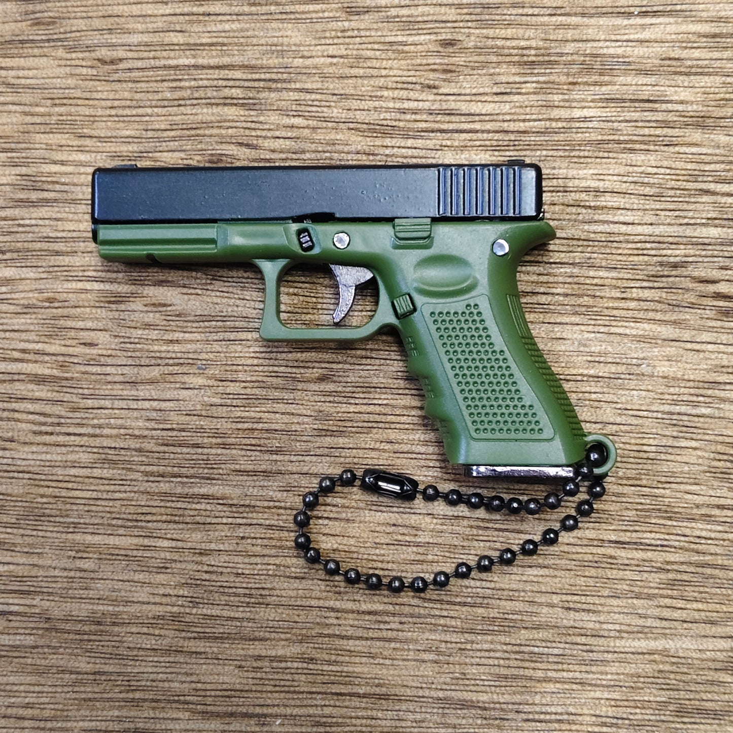 A compact pistol-shaped keychain or toy in olive green and black, shown from the side on a wood-grain surface with a black bead chain attached.