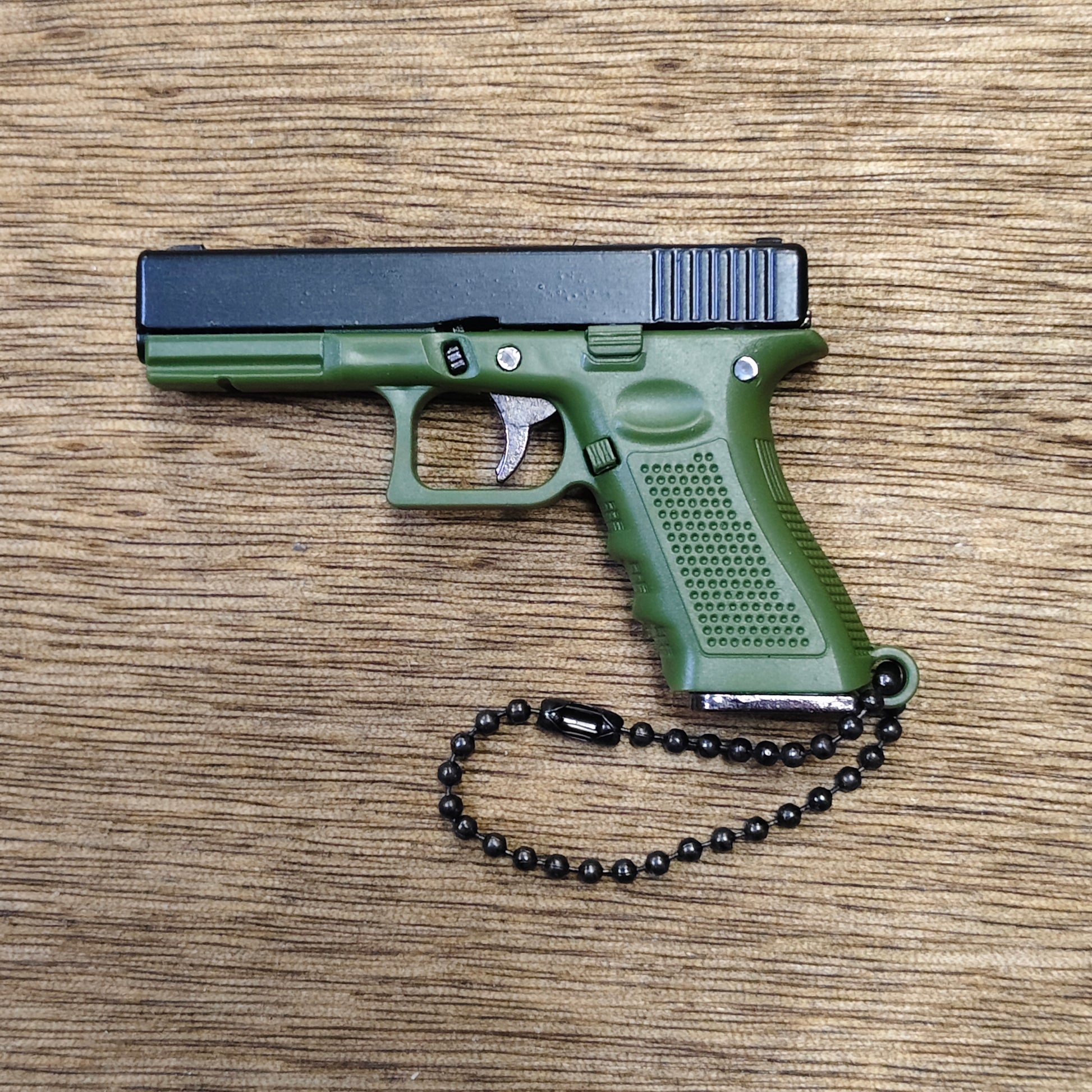 A compact pistol-shaped keychain or toy in olive green and black, shown from the side on a wood-grain surface with a black bead chain attached.