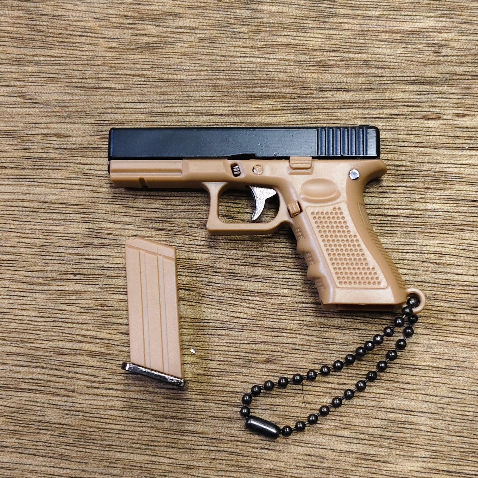 A toy semi-automatic pistol keyring in tan and black, shown with a matching tan magazine and a black bead chain on a wood-grain surface.