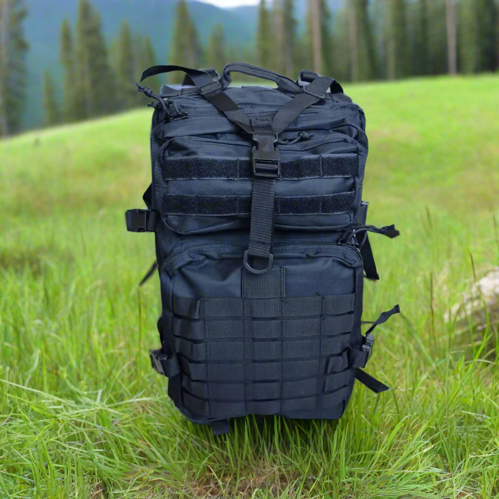 Black backpack surrounded by green grass, in a forest, mountains, and trees in the background